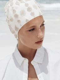 Model wearing a beaded white swim cap and white collared shirt against a bright sandy beach background