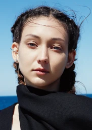 Close-up portrait of a woman with braided dark hair wearing black attire against a bright blue sky and ocean background.