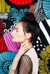 Profile portrait of a woman with an elaborate topknot hairstyle, winged eyeliner, and layered colourful bangles against a vibrant patterned backdrop