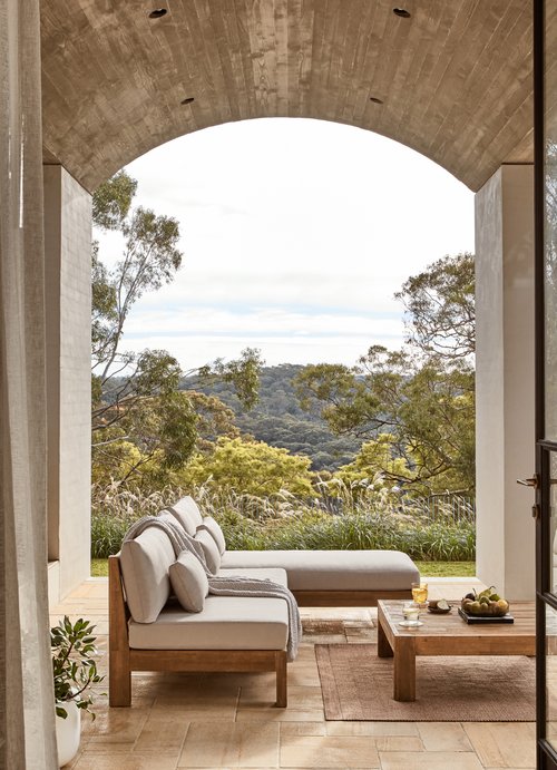Outdoor living area features a wooden sofa with cushions, a coffee table with fruit, and a scenic view of trees and hills.