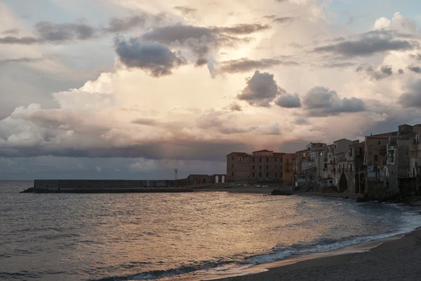 Coastal Italian village scene at sunset with historic buildings, tranquil ocean waves, and dramatic cloudy sky with soft light.