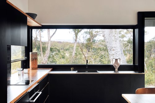 Sleek kitchen interior with black cabinets, light wood countertop, minimalist decor, and a large window showcasing a vibrant forest view.