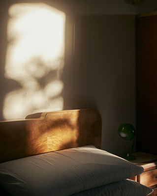 Bedroom interior featuring a bed with white pillows and a wooden headboard, illuminated by soft sunlight with a green desk lamp.