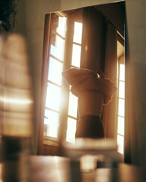 Reflection of person in a mirror with window light shining. Interior shot of figure adjusting clothing with bright daylight.