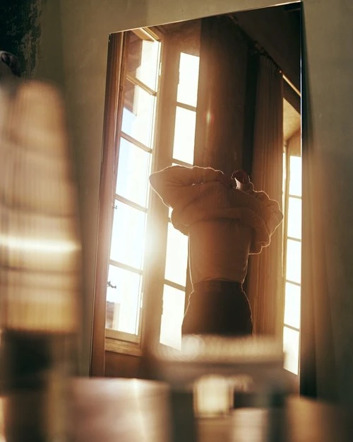 Reflection of person in a mirror with window light shining. Interior shot of figure adjusting clothing with bright daylight.