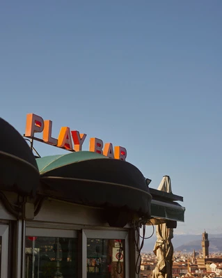 Outdoor shot of the PLAY BAR sign above a dark green awning and white trim, set against a blue sky in Florence, Italy.