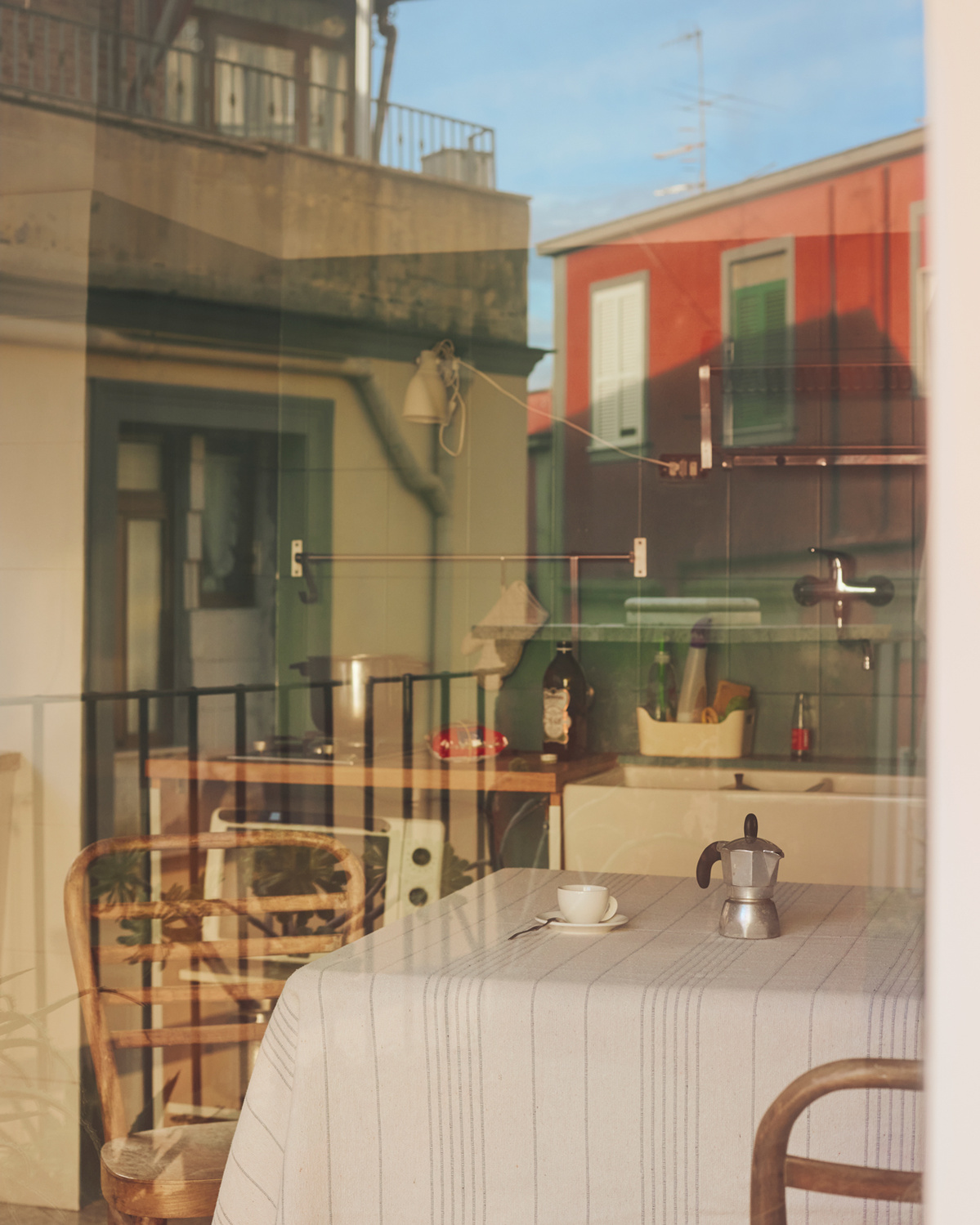 Still life indoors featuring a breakfast table set with coffee cup and moka pot, visible through a window with a building reflection.