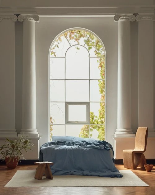 Interior with a blue bed placed in front of a large arched window, flanked by white columns, along with a chair and plants.