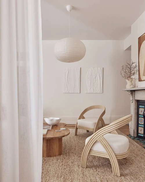 Neutral living room with two woven armchairs, a round wooden coffee table, jute rug, white pendant light and modern wall art.