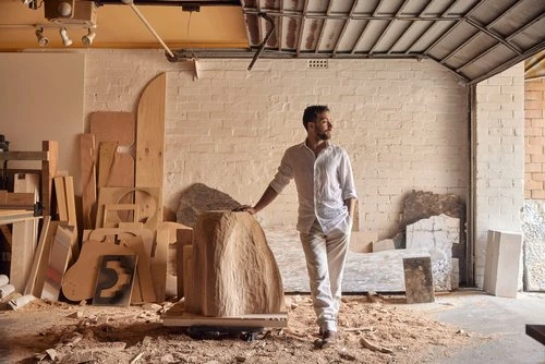 Bearded man in a white shirt and pants leaning on a carved sculpture in a wood-filled artist studio with overhead lighting and open doorway.
