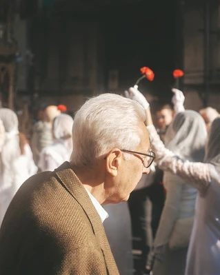 Elderly man in glasses and a brown jacket is the primary subject, with a crowd of people holding red flowers in the background.