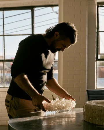 Man with beard and dark shirt examining a crystal cluster on a table inside a light-filled studio by Nic Gossage