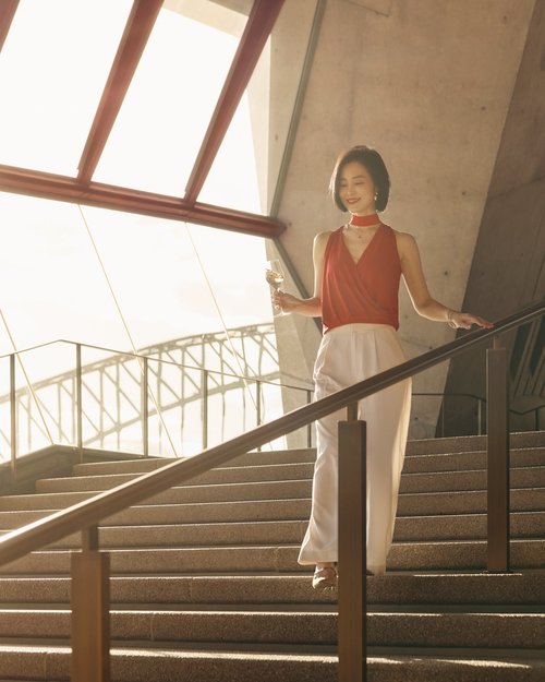 Woman in a red top and white pants descends the stairs of an urban building, holding a glass with bright sunlight in the background.