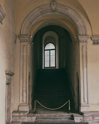 Architectural view of a stone staircase beneath an archway and a window at the top of the stairs, with a rope barrier.