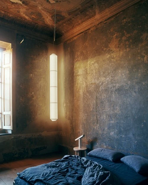 Interior view of a bedroom with a hanging lamp, blue bed, and textured walls providing a moody atmosphere and soft, diffused light.