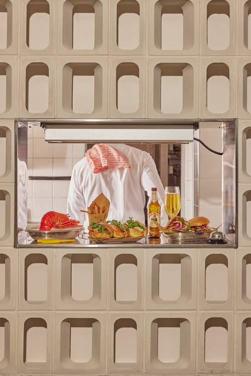 Food service featuring a chef behind a serving window presenting a lobster, fries, sandwiches, beer, and a hamburger on trays.