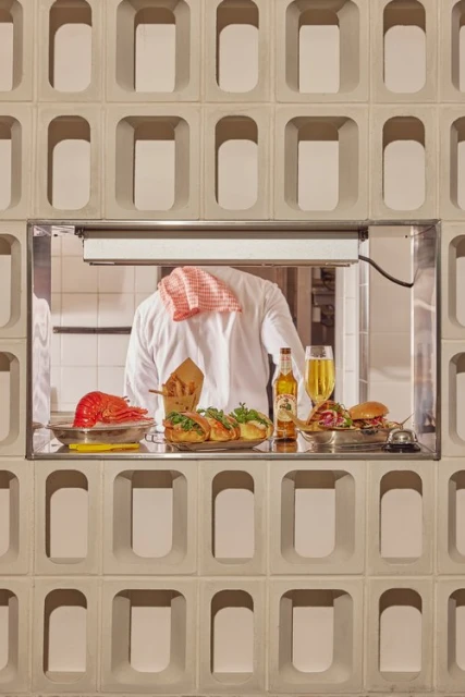 Food service featuring a chef behind a serving window presenting a lobster, fries, sandwiches, beer, and a hamburger on trays.