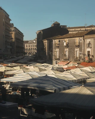 Aerial view of outdoor market stalls under blue and white striped canopies in a European city square with surrounding buildings.