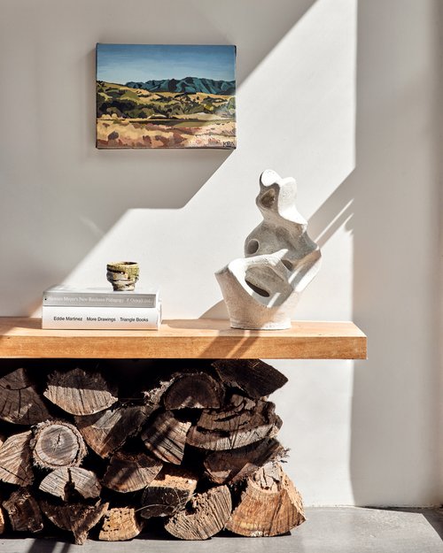 Contemporary interior featuring a painting of a landscape, a modern abstract sculpture, books, firewood, and sunlight casting shadows on a white wall.