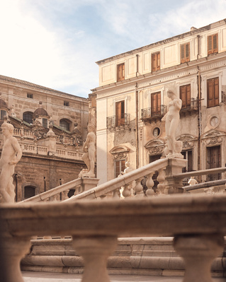 Cityscape featuring statues atop stone staircase in front of European-style buildings with balconies and windows under a bright sky.