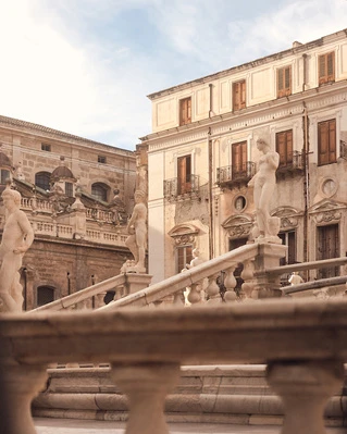 Cityscape featuring statues atop stone staircase in front of European-style buildings with balconies and windows under a bright sky.