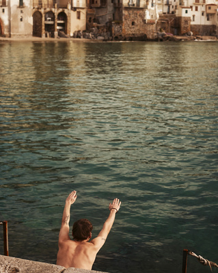 Back view of a shirtless man with arms raised, standing by the sea with buildings visible in the background. Travel photography.