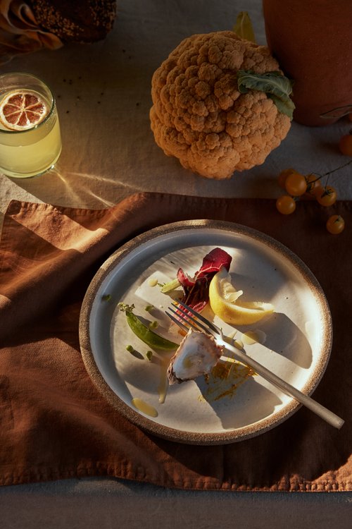 Overhead shot of a plate with meal remnants, a glass of beverage, and a cauliflower on a linen-covered table with a brown napkin.