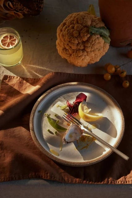 Overhead shot of a plate with meal remnants, a glass of beverage, and a cauliflower on a linen-covered table with a brown napkin.