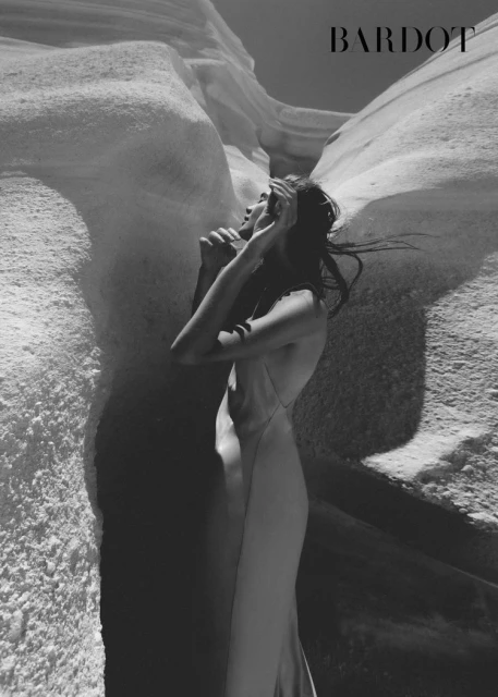 Woman in flowing gown poses in desert canyon landscape with white rock formations during a fashion shoot, artistic monochrome.