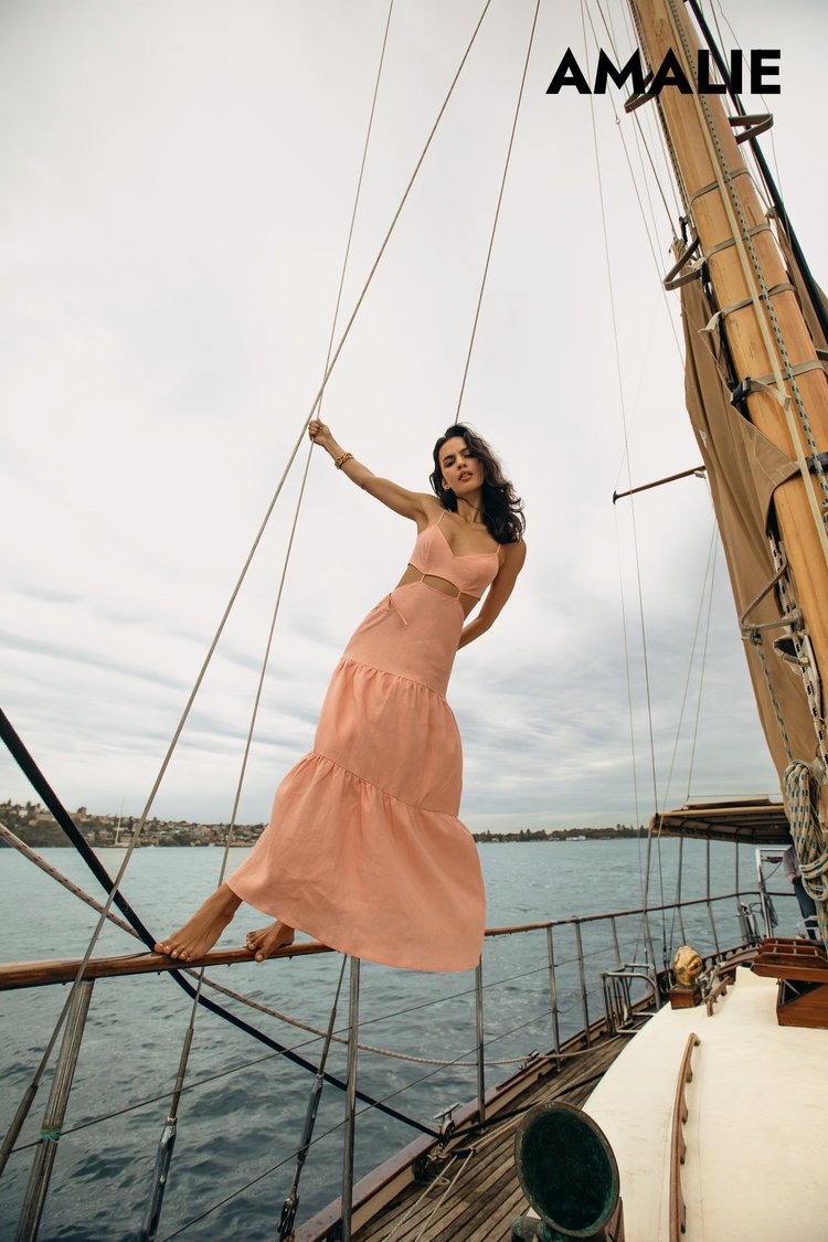 A woman wearing a peach dress poses barefoot on a sailboat with water and cloudy skies visible in the background for a fashion shoot.