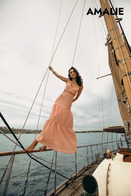 A woman wearing a peach dress poses barefoot on a sailboat with water and cloudy skies visible in the background for a fashion shoot.