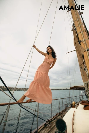 A woman wearing a peach dress poses barefoot on a sailboat with water and cloudy skies visible in the background for a fashion shoot.