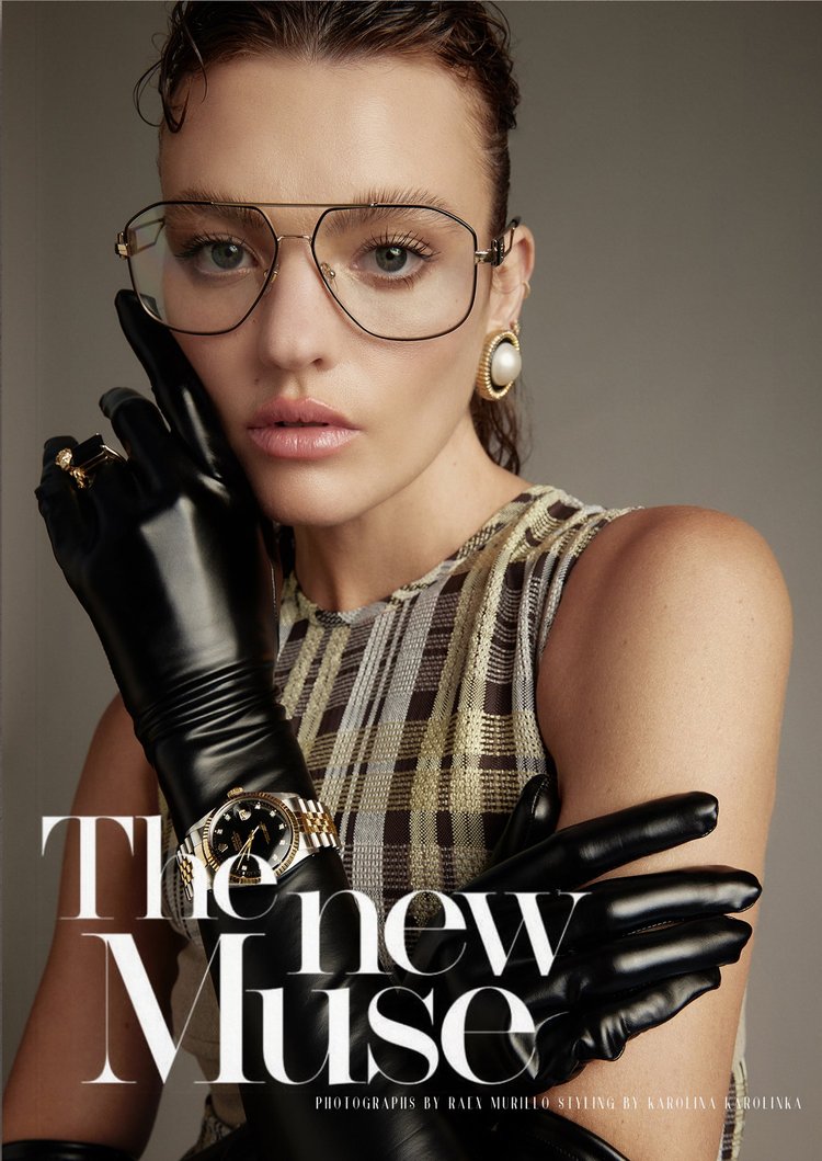 Fashion editorial shot of a woman wearing glasses, a plaid top, black gloves and gold jewelry in a studio with a neutral background.