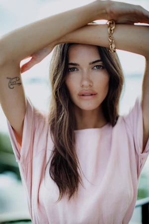 Portrait of a woman in a pink top with arms raised, wearing a gold chain bracelet and featuring a small tattoo on her upper arm.