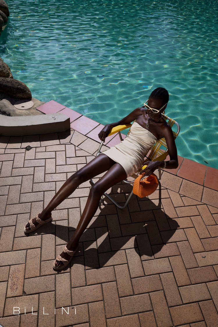 Woman in dress relaxes on a chair near a turquoise swimming pool, wearing sunglasses and sandals, holding an orange purse.