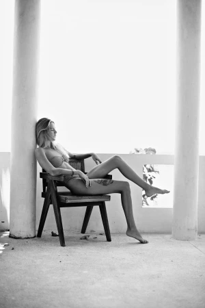 Woman in bikini seated on a dark wooden chair between white architectural columns, photographed in black and white by Raex Murillo.