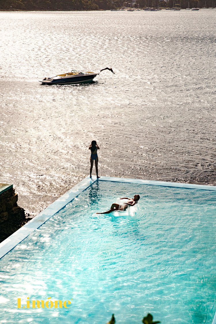 Woman on inflatable raft in pool, with another woman standing on pool edge overlooking a speedboat in the ocean under sunlight.