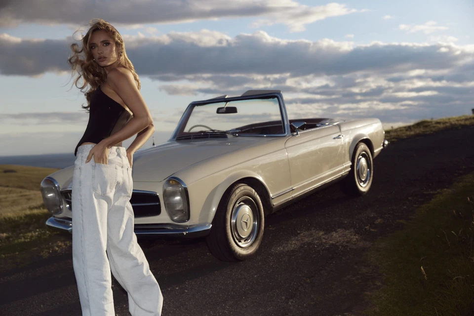 Blonde woman in a black top and light jeans poses near a vintage cream convertible car on a road against a cloudy sky.