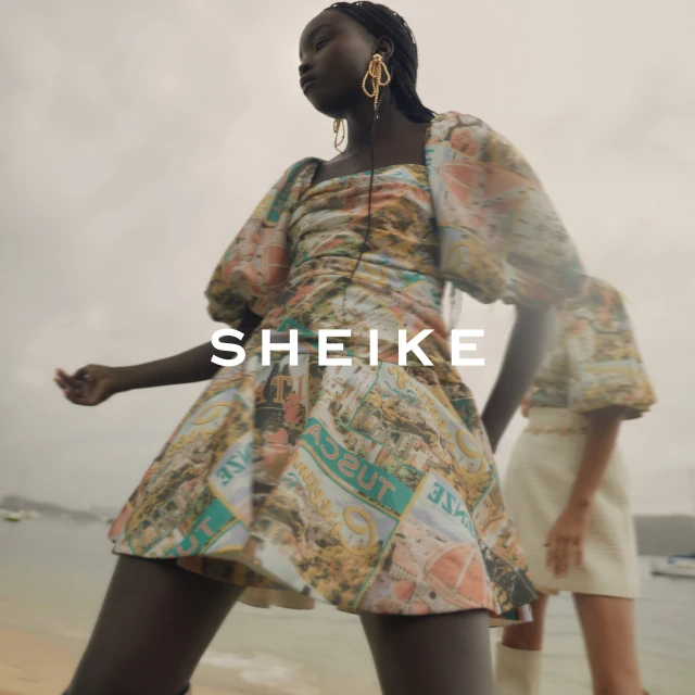 A woman in patterned mini dress poses on a beach near the ocean. Another woman is partially visible behind her on the sandy shore.