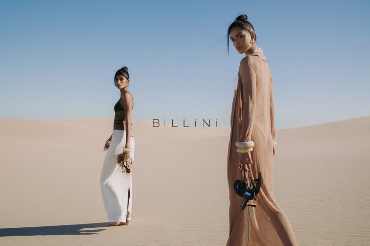 Two women in flowing dresses stand on a sand dune in a desert landscape. The woman on the right carries sandals in her hand.
