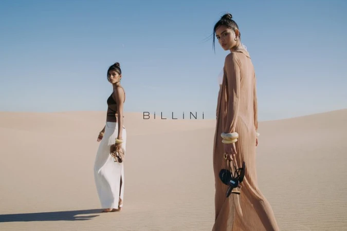 Two models walking across sand dunes in a desert landscape carrying sandals under a clear blue sky