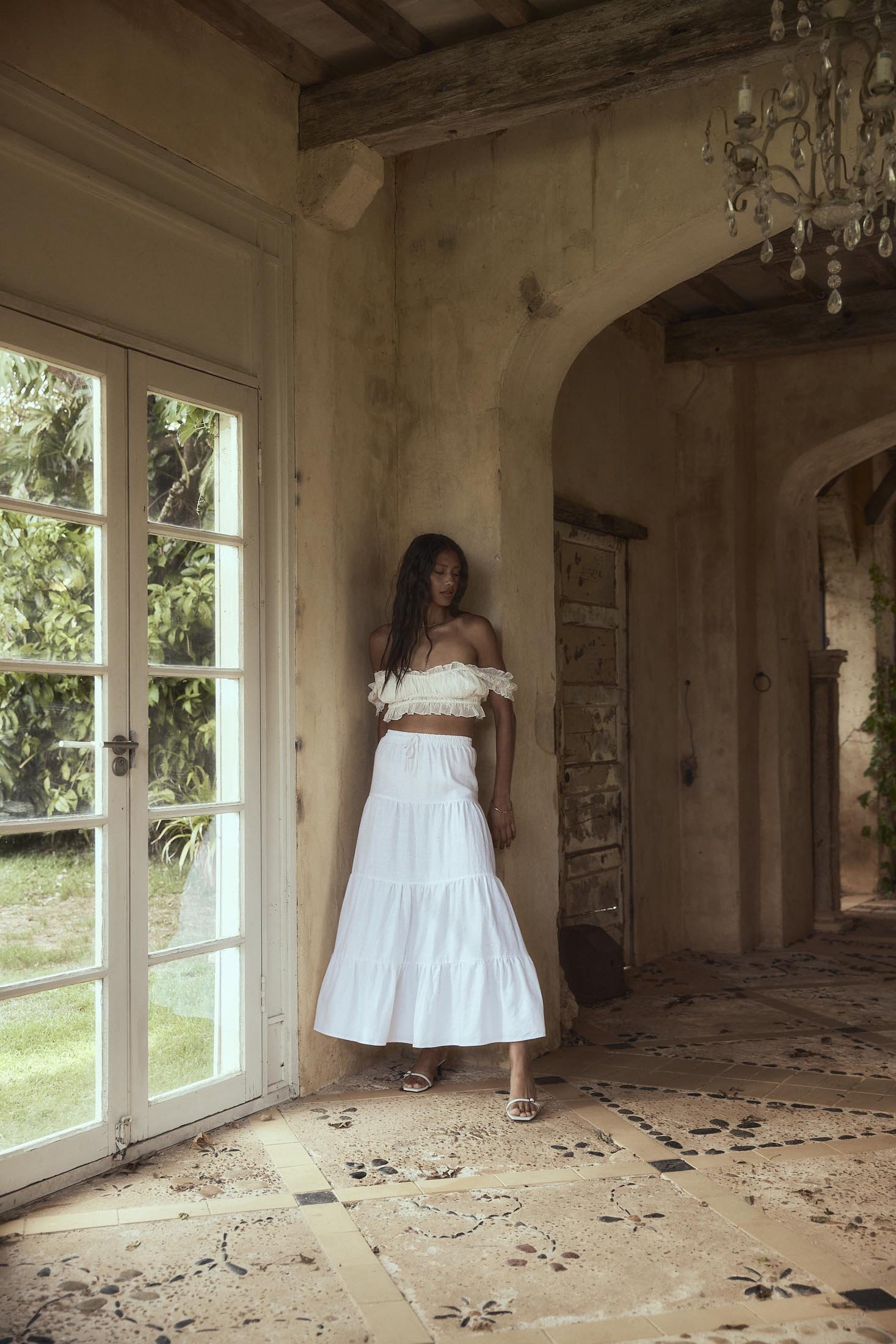 Young woman poses in a ruffled white top and long skirt inside an old building with stone tile floor near a doorway and windows.