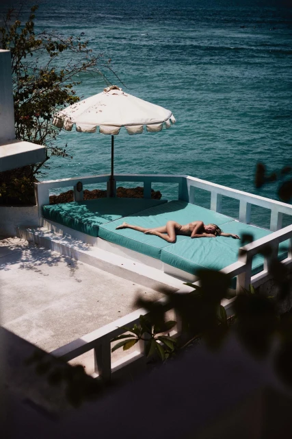 Adult female sunbathing on a teal lounger under a white umbrella with the ocean visible in the background on a sunny day at the resort.
