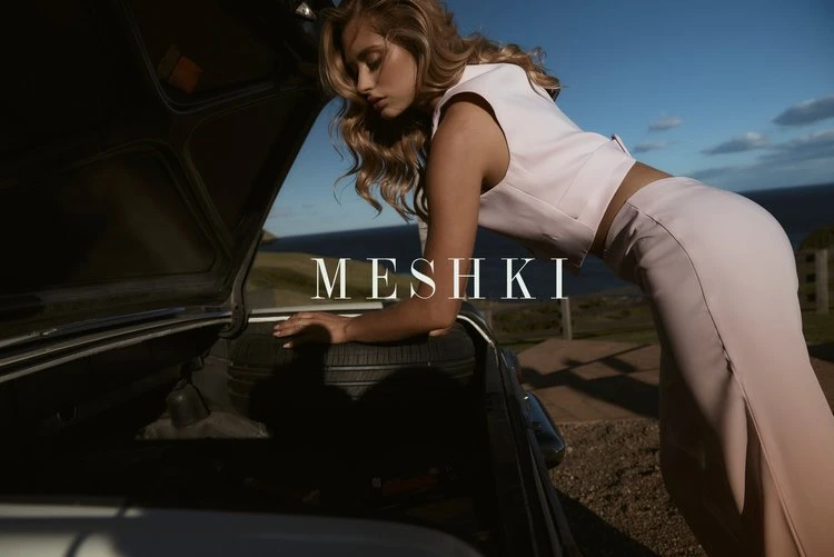 A woman in a pale pink two-piece outfit poses by the open trunk of a car with a tire inside against a backdrop of ocean and sky.