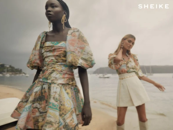 Two female fashion models pose on a beach wearing dresses with vintage prints during daytime, showcasing summer collection.