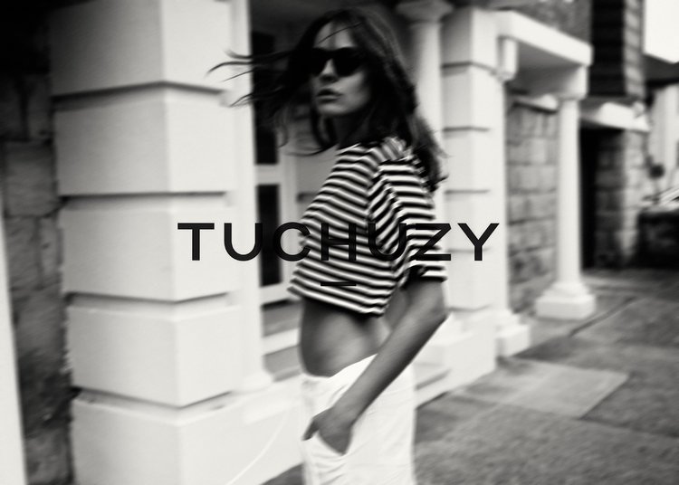 Woman in striped crop top and white pants poses near a building. Fashion shot features black and white tones, sunglasses, and streetwear.