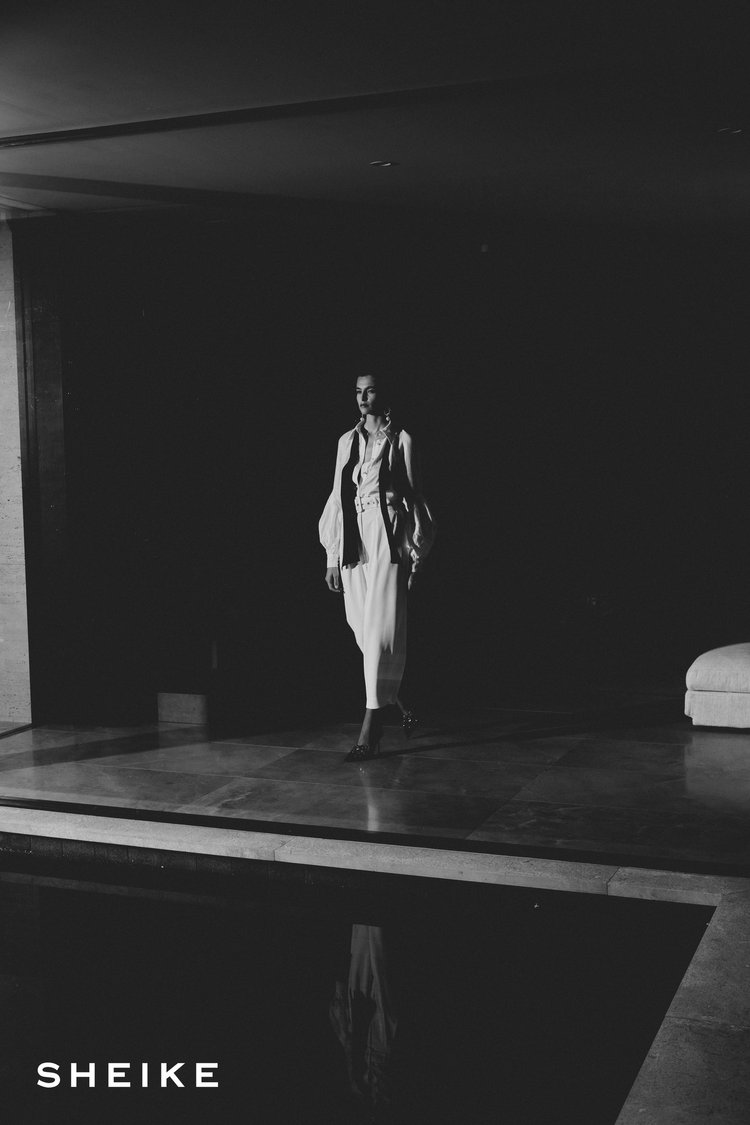 Elegant portrait of a female fashion model in white dress walking indoors in a dark room with reflective pool, and designer logo.