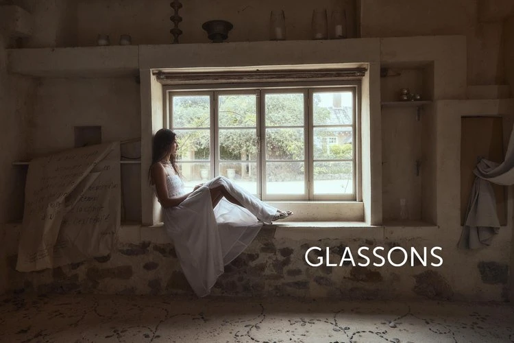 Woman in a white dress sits in a window looking out at greenery in a sunlit indoor setting, creating a serene and dreamy atmosphere.