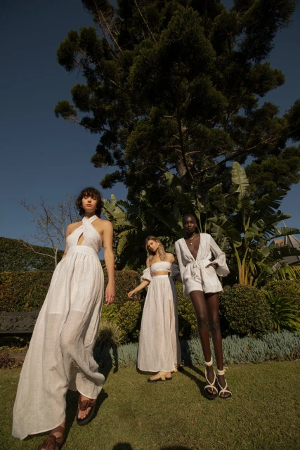 Three women model white dresses in an outdoor fashion shoot, standing on a grassy lawn with lush green foliage and a blue sky backdrop.