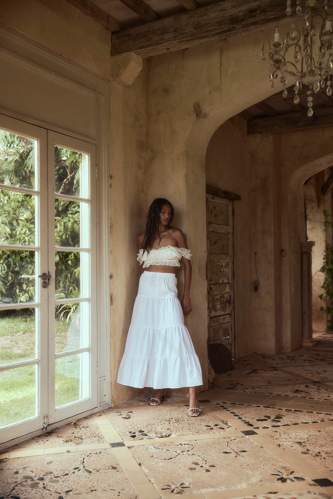 Woman wearing a white off-the-shoulder crop top and long white tiered skirt standing in an old stone hallway by a large glass door.